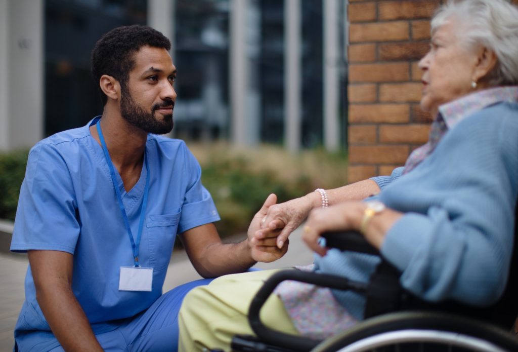 Betterdays Healthcare A healthcare professional in blue scrubs attentively listens to an elderly woman in a wheelchair during an outdoor conversation.